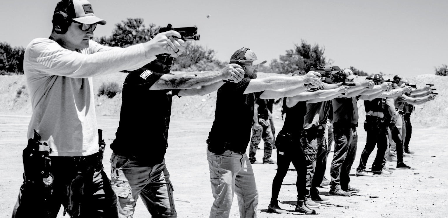 Large group of tactical students training at the range, firing pistols in a synchronized line under instructor supervision